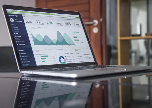 A silver laptop sits on a reflective black surface, displaying a business analytics dashboard with various graphs, charts, and performance metrics. 